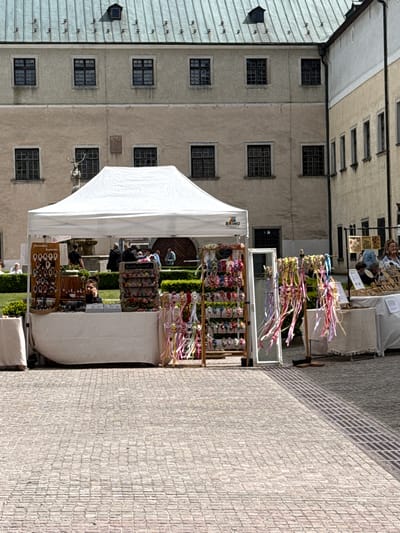 An outdoor market or craft fair is depicted in a charming European town setting, likely in Častá, Slovakia. The scene is bathed in bright daylight under clear skies.
In the mid-ground, a prominent white tent, displaying the brand "BRIMO" on its side, serves as a stall. Beneath and around it, various artisanal products are showcased. Notable items include numerous colorful floral headbands and wreaths adorned with long ribbons, some hanging on a display rack and others neatly arranged on shelves. Further inside the tent, a display of what appears to be handmade jewelry, possibly bracelets and necklaces, is visible, with price tags or labels displaying numbers like "00000" and "500". A person's face is clearly visible peeking out from behind this jewelry stall.
Several other market stalls are scattered in the background, with people browsing or working. One sign in the background reads "Langosaren," suggesting food sales. Another visible text on a stall appears to be "Nirodaé hamene."
The market is situated on a cobblestone plaza or courtyard. Surrounding the market are tall, light-colored stone buildings with numerous dark, rectangular windows, typical of older European architecture. To the far left, a building with a grey corrugated metal roof and multiple small, dark openings is visible. One building also features a decorative architectural element resembling antlers or a deer head sculpture. The overall atmosphere appears calm and inviting, characteristic of a local daytime market event.