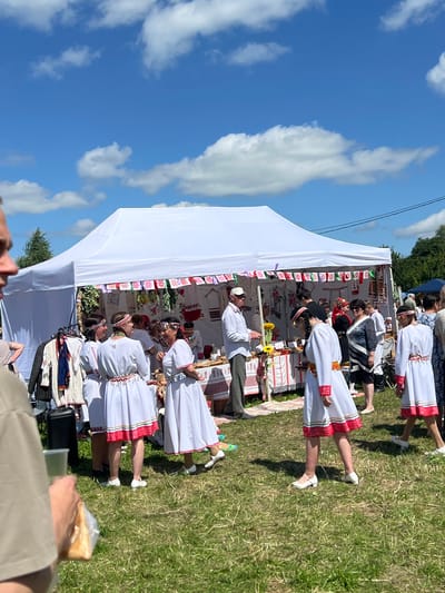 The image depicts an outdoor gathering under a clear blue sky with scattered white clouds, likely at a festival or market in Kalinino, Russia. A large white tent serves as a central structure, decorated with colorful, folkloric banners. Inside and around the tent, people are engaged in various activities.
Several individuals, predominantly women, are dressed in traditional white outfits adorned with red and patterned embroidery. Some are standing and conversing, while others appear to be interacting with a stall. One woman in a white dress with red trim is holding her hands up in a gesture of explanation or demonstration. Another group of women in similar attire are gathered, looking towards the center of the scene.
In front of the tent, a table is set up with various items, suggesting a sale or display of goods. These items are not clearly identifiable but appear to be small and varied. To the left of the tent, clothing items are displayed on a rack, including jackets and shirts with intricate embroidery.
A man in a white shirt and light-colored pants, wearing a cap, stands near the table, possibly attending to customers or presenting items. Another man, partially visible in the foreground and out of focus, is holding a clear plastic cup and a bag containing what appears to be food.
The ground is covered in green grass, indicating a natural or park-like setting. The overall atmosphere seems lively and festive, with a strong emphasis on cultural tradition, as evidenced by the attire and decorations.