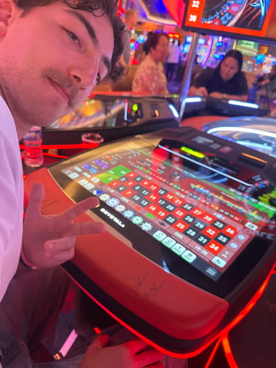 Here is a concise description of the image:
The image appears to be a selfie taken by a person with dark hair and a mustache, posing at a digital roulette table. The table is orange with a black trim and has a screen displaying the roulette layout and betting options. The screen displays the numbers 1-36, as well as 0, 00, and various betting options like "Even", "Odd", "1-2", "19-36". In the background, other casino patrons and gaming machines are visible, suggesting that the scene is in a casino. The logo "CRYSTALE" is visible on the table. The person in the foreground is making a peace sign.
Given the presence of roulette machines and casino environment, it's likely this photo was taken at the Seminole Hard Rock Hotel & Casino in Hollywood, Seminole Reservation, United States of America.