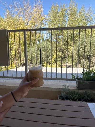The image displays an outdoor scene from a balcony or patio in the city of Maale Adumim, Palestinian Territory, under clear daylight. In the foreground, a light-skinned right hand with a thin black bracelet on the wrist holds a clear, ribbed glass filled with an iced, light brown beverage topped with white foam and a black straw. The hand and drink are positioned above a dark brown slatted table that occupies the bottom third of the frame. To the right, on a light beige tiled ledge, several potted plants are visible: a rectangular dark gray planter containing tall green herbs, and three smaller square pots holding varied green and grayish-green foliage. A gray metal railing with vertical bars runs across the midground. Beyond the railing, a portion of a corrugated metal roof is visible. The background consists of numerous trees with predominantly yellow-green leaves, set against a bright blue sky.