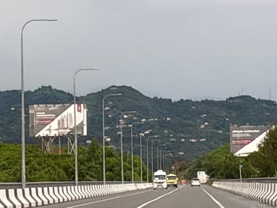 The image shows a view from a highway in Chakvi, Georgia, with lush green hillsides in the background. The highway is lined with streetlights and a concrete barrier with black and white stripes. Several vehicles are visible on the road, including a white truck and a yellow van. Billboards are placed on either side of the highway, with one on the left displaying an advertisement in Georgian script and imagery of architectural elements, and another on the right with similar content. The sky is overcast, suggesting a cloudy day. The terrain is hilly, with houses scattered across the slopes. The overall scene depicts a travel route through a scenic, semi-urbanized coastal area.