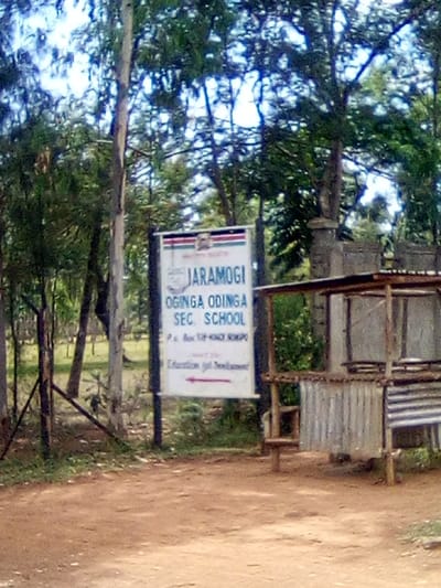 The image displays the entrance area of Jaramogi Oginga Odinga Sec. School, located in West Sakwa ward, Bondo, Siaya County, Kenya. A white rectangular sign, centrally positioned, bears the school's name "JARAMOGI OGINGA ODINGA SEC. SCHOOL" in blue text, along with the Ministry of Education logo and Kenyan flag at the top. Below the name, "P.o. Box 514-BONDO" is printed, and at the bottom, "Education for Development" with a red arrow pointing left. To the right of the sign, a rustic, open-sided structure composed of wooden poles and corrugated metal sheets is visible. The foreground consists of reddish-brown dirt. The background is dominated by tall trees with green foliage, partially obscuring the sky. No individuals are present.