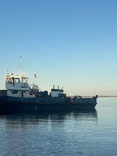 The image depicts a barge on a body of water, likely a river or lake, with Soroch'i Gory, Russia, in the background. The barge is dark blue and white, with a multi-level structure including a pilot house, antennas, and a Russian flag. The water is calm and reflects the barge. The sky is a clear, light blue. A strip of land is visible in the distance.