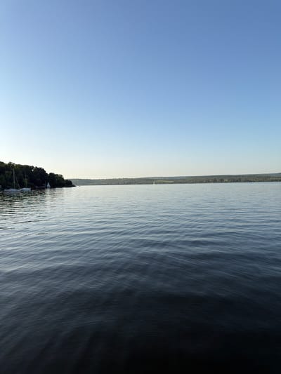 This is a landscape photograph taken on a clear day, likely in the late afternoon or early evening, given the warm, soft light. The image is oriented vertically. The dominant subject is a large body of water, likely a lake or a wide river, which occupies the right two-thirds of the frame. The water's surface is textured with gentle ripples that catch the light, creating a dynamic pattern of light and shadow.
The left third of the image is a vast expanse of clear blue sky, with a slight gradient towards a lighter hue near the horizon. In the distance, along the far edge of the water, is a thin strip of land densely covered with trees, suggesting a natural, possibly forested coastline. A few faint structures or buildings can be discerned within the treeline, hinting at a settlement. A single white sailboat is visible on the water, a small speck against the immensity of the lake and sky. The overall mood is serene and peaceful, with a sense of calm and quietude. The location is identified as Vatra, Moldova. No text is visible in the image.
