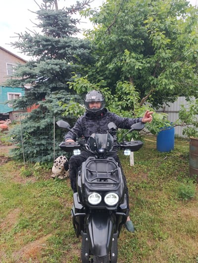 In the city of Novoye, Russia, a man in a black camouflage jacket and helmet sits on a black all-terrain vehicle. He is facing forward, with his right hand raised, index finger pointing. The ATV has two prominent headlights and a cargo rack at the front. Beside the ATV, a stuffed dog toy with black spots lies on the grassy ground. Behind the ATV, a large coniferous tree stands prominently, with a wooden house and a metal fence visible in the background. Green foliage from trees and bushes surrounds the scene. A large blue plastic barrel is also situated behind the ATV. The overall environment suggests an outdoor, possibly rural or suburban setting.