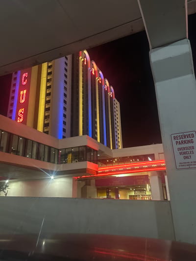 The image displays the exterior of the Circus Circus hotel-casino in Reno, Nevada, United States of America, during nighttime. The perspective is from a lower level, likely a parking structure or covered driveway, looking upwards. The main hotel tower features a facade illuminated with vertical bands of yellow and blue light. On the left side of the tower, large red neon letters "C," "U," and "S" are vertically stacked, forming part of the "CIRCUS" signage. Arched neon elements are visible on the right side of the building's upper section.

Below the main tower, an elevated, glass-enclosed pedestrian walkway, internally lit, spans horizontally across the mid-ground. Beneath this, a structure with a prominent red neon strip marks a covered drive-through or entrance area. Signs in this area indicate "LUGGAGE DROP OFF," "THRU TRAFFIC," and "CIRCUS CIRCUS HOTEL REGISTRATION."

In the immediate foreground, a light-colored concrete barrier or wall runs across the bottom of the frame. On the far right, a substantial, light-colored concrete column or support structure is present, extending vertically. A white and red sign affixed to this column reads: "RESERVED PARKING OVERSIZED VEHICLES ONLY," followed by "VIOLATORS WILL BE TOWED AT VEHICLE OWNERS EXPENSE." An overhead concrete beam partially obscures the top portion of the main building. Multiple artificial light sources generate visible lens flares in the lower half of the image. No individuals or specific actions are discernible within the scene.