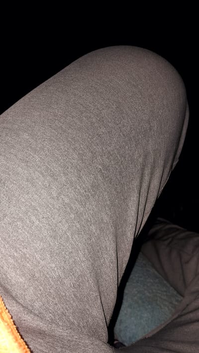 The image displays a close-up view of a person's upper leg, likely a thigh, covered in light grey marled fabric, consistent with sweatpants or casual trousers. The fabric shows a distinct woven or knit texture. The leg appears rounded, suggesting the person is seated or resting.
In the upper right portion of the frame, a darker, possibly olive-green or brown, fabric is visible, which could be part of another garment or a blanket. Beneath this darker fabric and further in the background, a patch of textured, bluish-green fabric, resembling fleece or a soft upholstery, is present, indicating a cushioned surface like a seat or bed. A small fragment of vibrant orange or yellow fabric is just visible at the very bottom right edge.
The scene is characterized by very low lighting conditions, casting most of the surroundings into deep shadows, which suggests an indoor setting or nighttime. Thin, dark lines, possibly seams or elastic bands, run across the grey fabric. No specific activity is taking place other than the person's leg being in this position. There are no discernible location cues to pinpoint the setting to Umuigbekwo, Nigeria, and no visible text is present.