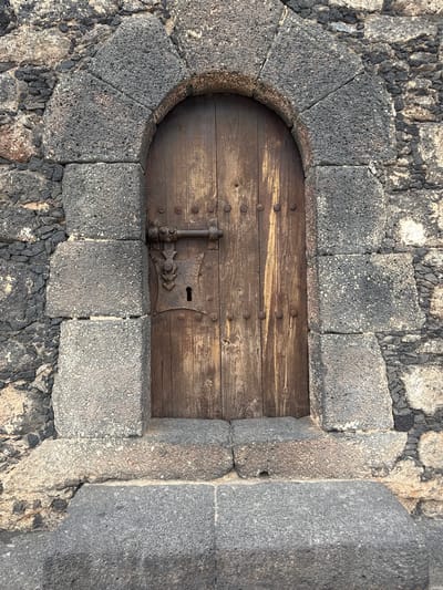 A high-angle, medium shot depicts a small, closed wooden door, likely a cellar or storage entrance, set into a stone structure, potentially a wall or foundation. The door is made of horizontal wooden planks, secured with metal studs and featuring a simple, aged metal latch and keyhole plate. The surrounding stonework consists of rough, dark gray, porous stones, some cut into geometric shapes to form an arched doorway. The stone blocks are fitted together with small dark stones. The overall setting suggests an old building or fortification.

Based on visual characteristics, the location is likely in Arrecife, Spain, which has a variety of architecture using dark volcanic stone.