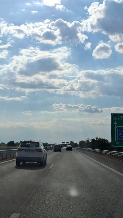 The image depicts a highway scene in Bulgaria. A white SUV, with license plate CB 8379 TC, is driving on the right lane of the highway. Several other vehicles are visible ahead in the distance, traveling in the same direction. A green highway sign is on the right side of the road, indicating directions to Sofia and Tsalapitsa. The sky is partly cloudy with the sun shining through. The environment appears to be rural, with trees lining the sides of the highway.