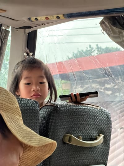 The image, taken inside what appears to be a bus or van, shows a young East Asian girl looking down at a smartphone she is holding. She has her hair pulled back in a bun with a butterfly clip. The girl is seated behind a textured dark grey headrest. In the foreground, the brim of a straw hat is visible, obscuring the face of another person. The background shows a window with water droplets on it, indicating it may be raining, and through the window, glimpses of trees and a red roof are visible. The interior of the vehicle appears to be made of grey fabric and plastic. The location is identified as Palai, Laos.