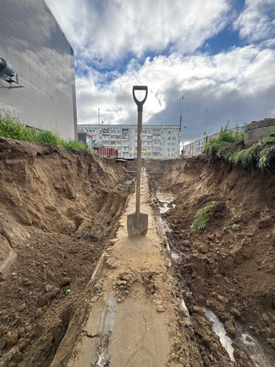 The image shows a deep excavation, likely a trench, in an urban setting. A shovel with a black D-shaped handle is partially embedded in the loose soil at the bottom of the trench, suggesting recent digging activity. The walls of the trench are rough and earthen, with some signs of water or mud. In the background, a multi-story apartment building with numerous windows is visible, indicating a residential area. A modern building with a reflective facade is also present in the foreground to the left. Overhead, the sky is partly cloudy with patches of blue sky and bright white clouds. Utility poles and wires are visible between the excavation and the apartment building. The overall impression is of ongoing construction or maintenance work in a built-up area. The lighting suggests it is daytime, likely overcast with some sun breaking through. The location context of Usinsk, Russia, places this scene in a northern climate, which could influence the type of construction and the appearance of the soil. There is no visible text in the image.