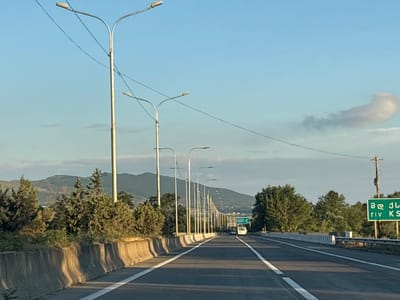 This image depicts a highway scene in Mukhrani, Georgia. The perspective is from a vehicle traveling on the road, looking forward. A series of streetlights line the right side of the highway, with their lights off, suggesting it is daytime. The sky is a clear blue with a few wispy clouds. On the left side of the road, a concrete barrier separates the highway from a landscape of trees and rolling hills. A green road sign with text in Georgian and English is visible on the right side of the highway, indicating directions to "riv. KS". Two vehicles, a white van and a darker car, are visible further down the road. The overall environment appears to be rural or semi-rural, consistent with a highway outside of a major urban center.