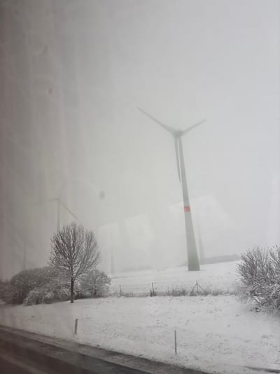 This image captures a snow-covered rural landscape in Schöpstal, Germany, enveloped in dense fog or mist. In the mid-right, a prominent wind turbine with a grey tower featuring a red stripe and three blades stands partially obscured by the atmospheric conditions. A second wind turbine is faintly visible in the left-center background, heavily veiled by the fog. The ground is uniformly covered in white snow. A dark, wet surface, resembling an asphalt road, occupies the immediate foreground. To the left, a leafless deciduous tree and several snow-dusted bushes are situated behind a wire fence with dark posts. The sky is light grey and overcast. Reflections consistent with an interior window are present across the image surface, indicating the photograph was taken from within an enclosed space. No individuals, animals, or active subjects are present, and no discernible actions or interactions are depicted.