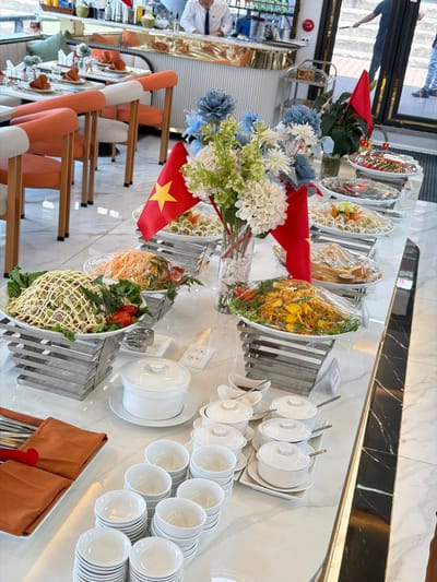 This indoor scene captures a meticulously arranged buffet spread, likely within a hotel or restaurant setting in Tuan Chau Ward, Vietnam. The central feature is a long, gleaming white buffet table with a marble-like surface, showcasing a variety of dishes.

Numerous oval-shaped serving platters hold different salads and prepared food items, many covered with clear plastic wrap to preserve freshness. These dishes display colorful ingredients such as shredded carrots, green leafy vegetables, and other mixed components. Smaller, individual portions, possibly spring rolls, are also visible.

Adjacent to the food, stacks of clean, white ceramic bowls, plates, and lidded serving tureens are neatly organized for guest use. Cutlery, including spoons and forks, is presented in tidy burnt orange fabric pouches.

Decorative elements include elegant artificial floral arrangements featuring blue, white, and green flowers in clear vases. Small Vietnamese flags, identifiable by their red color and yellow star, are strategically placed throughout the buffet, subtly indicating the location.

In the background, to the left, a dining area with orange and white upholstered chairs and tables is partially visible, hinting at the seating arrangements. A staff member in a white shirt can be seen attending to the area, and a reflection of a person's lower body is visible in a polished surface or window, suggesting an active environment.

The overall presentation suggests a formal or semi-formal dining experience, with the buffet prepared for a meal service. The scene is orderly and well-lit, though the specific time of day is not discernible. No prominent text is visible on any signs or labels.