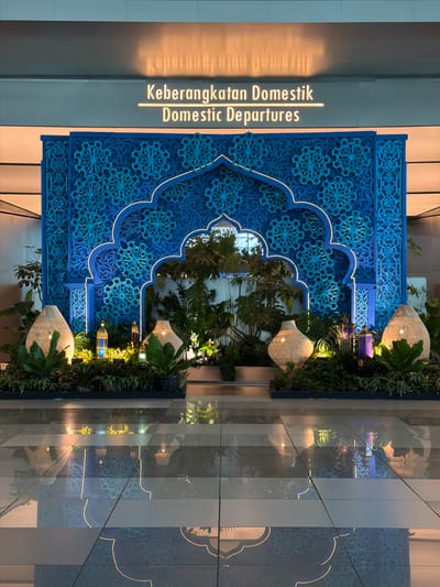 The image displays an ornate indoor setting, likely within an airport terminal in Pajang, Indonesia, designated for domestic departures. A prominent feature is a large, rectangular blue decorative screen, positioned vertically, adorned with intricate, symmetrical floral and geometric patterns typical of Islamic or Indonesian artistry. The central arch-shaped cutout and the upper frame of this screen are outlined with a warm, glowing light.
To the left of the blue screen, against a neutral-toned wall, illuminated text is visible. It clearly states "Keberangkatan Domestik" above "Domestic Departures," identifying the area's function. The text itself emits a soft, golden light.
Immediately adjacent to the blue panel on the right is a vibrant vertical garden, densely planted with various green foliage. Embedded within this greenery are several decorative lanterns or light fixtures, crafted from a light-colored, woven material. These lanterns glow with a warm yellow light, illuminating the plants. Smaller, colorful glass light fixtures are also interspersed among the foliage.
The lower part of the scene features a highly reflective floor or surface, creating clear, inverted reflections of the blue screen's patterns, its illuminated outline, the green plants, and the glowing lanterns. The overall impression is of an elegant, culturally rich environment, likely viewed during the evening or under artificial light due to the prominent use of warm illumination. There are no people visible, and no specific activities are taking place; the focus is on the decorative architecture and lighting.