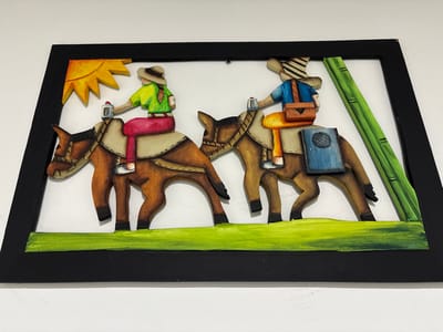 This image displays a framed, three-dimensional wooden relief artwork, characteristic of local craftsmanship in Quimbaya, Colombia. The scene depicts two stylized brown donkeys or mules, each with a light tan saddle, moving in profile from left to right across a horizontal strip of green representing ground. The upper left corner features a yellow, multi-rayed sun, while the upper right includes two vertical green stalks, resembling bamboo or sugar cane. The lead donkey is ridden by a female figure wearing a light brown hat, green shirt, and pink pants; her left hand holds a silver cylindrical object. The second donkey is ridden by a male figure wearing a striped hat, blue shirt, and light brown pants, carrying an orange satchel slung over his right shoulder. A dark blue, rectangular object with a circular textured pattern is affixed to the left side of the second donkey. The entire relief is mounted on a white background and enclosed within a plain black rectangular frame.