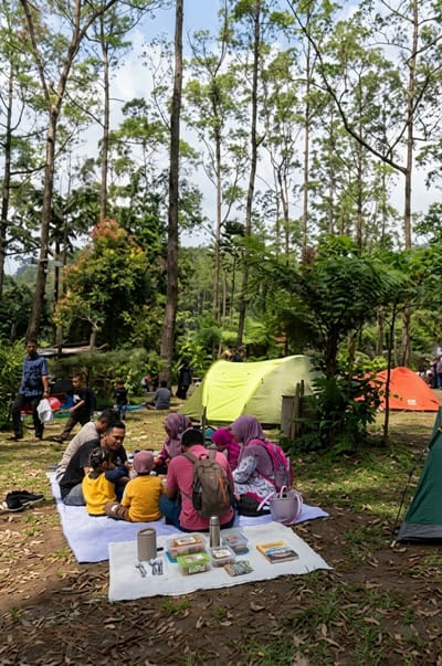 A group of people are picnicking on blankets laid out on the ground in a forest clearing. The scene takes place in Laugumba, Indonesia. Several tents are set up in the background, including a bright yellow one and an orange one. The people, some wearing headscarves, are seated in a circle around a picnic spread. They appear to be eating and socializing. A few children are also present. The surrounding environment is characterized by tall trees and lush green foliage, indicative of a forest. The ground is covered with dry leaves and grass. The lighting suggests it is daytime, with dappled sunlight filtering through the trees.