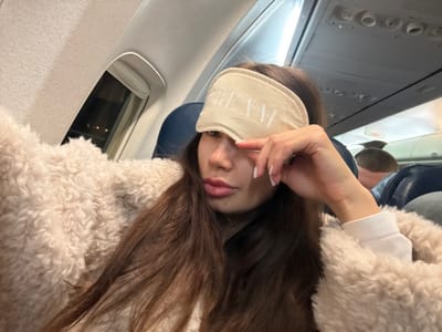 A woman with long brown hair wears a sleep mask with "DREAM" written on it, posed in what appears to be an airplane seat. She's wearing a fluffy cream-colored jacket. Her head is slightly tilted, and she has a hand near her face. A blue neck pillow is around her neck. An airplane window and the side of the plane interior are visible in the background, suggesting she is in flight.