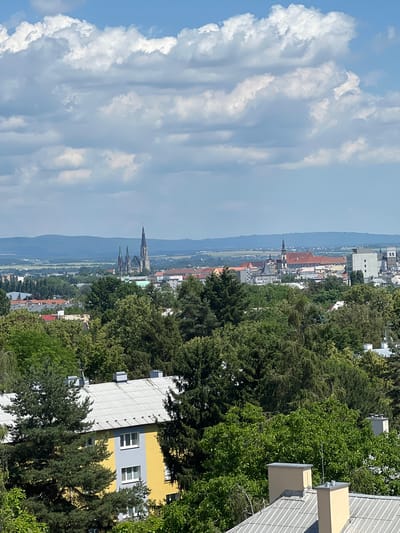 An aerial view captures a city skyline partially obscured by lush green trees. Dominating the background are the prominent spires of the St. Wenceslas Cathedral in Olomouc, Czechia. The cathedral's Gothic architecture, characterized by its tall, slender towers, stands out against the horizon. In front of the cathedral, various buildings with red-tiled roofs are visible, suggesting a historic city center. A modern apartment building, painted in yellow and gray, is situated in the foreground, with its white-tiled roof featuring several chimneys. The sky above is a vibrant blue, adorned with fluffy white clouds, indicating a clear and sunny day. The overall scene depicts a harmonious blend of natural landscape and urban development.
