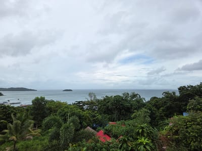 The image depicts a panoramic view of Koh Toch Village, Cambodia, on an overcast day. In the foreground, lush green foliage dominates, with palm trees and dense tropical vegetation. Several rooftops, some with red tiles, are visible peeking through the greenery. Beyond the foliage, the sea stretches out, a muted blue under the cloudy sky. The water appears calm, with a few small boats moored near a pier extending into the sea on the left side of the image. Further out, a small, tree-covered island is visible in the distance. The horizon is marked by a distant coastline with a low-lying landmass. The sky is filled with a blanket of grey and white clouds, with a few patches of pale blue sky showing through.