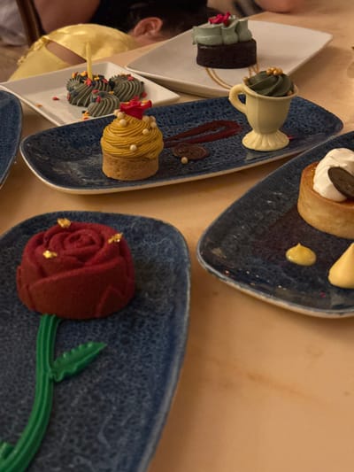The scene depicts a selection of themed desserts arranged on a light brown dining table within a restaurant setting. This location is identified as Be Our Guest Restaurant in Fantasyland, Magic Kingdom Park, Walt Disney World Resort, Bay Lake, Florida, United States.

In the foreground, to the lower left, a dark blue speckled rectangular plate holds a red, rose-shaped dessert with a textured, matte finish, adorned with small gold flakes on its petals. A sculpted green stem and leaf extend from its base onto the plate.

Adjacent and slightly further back, another dark blue speckled rectangular plate displays two primary desserts: a circular pastry base topped with a tall, swirling mound of mustard-yellow frosting decorated with small white spherical candies and a tiny red flower at its apex, and a white, molded ceramic cup resembling a teacup, containing dark olive-green frosting topped with gold sprinkles. Between these two items, a dark red berry coulis is artfully drizzled in a zigzag pattern, accompanied by small white and dark brown spherical garnishes.

To the upper right, a third dark blue speckled plate is partially visible, featuring a round tart or cheesecake topped with white whipped cream and a flat, dark circular chocolate garnish. Two small, pale yellow dollops of sauce are present on this plate.

Further into the background, on a white rectangular plate, multiple small, dark grey-green swirled frosting portions, likely on mini bases, are visible, some scattered with colorful sprinkles. A thin, lit yellow candle stands upright among these. Directly behind this, on a white plate, sits a small, dark chocolate brownie topped with light mint-green frosting and small pink rosette details.

In the blurred background, partially visible, are the upper torso and head of an individual wearing a yellow garment, possibly a character costume or a guest's attire. Another person's ear is also indistinctly visible. The overall impression is a festive dessert service, possibly celebrating an event given the presence of a lit candle.