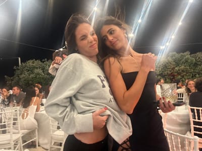 Two young women are posing for a photo at an outdoor evening event, likely a wedding reception, in Fasano, Italy. The woman on the left is wearing a light grey sweatshirt and black leggings, and is making a humorous expression with her eyes squeezed shut. She is holding the hem of her sweatshirt up, revealing her midriff. The woman on the right is wearing a black strapless dress and delicate gold jewelry, including a necklace and earrings. She is smiling slightly and looking towards the camera. The background shows a dimly lit setting with trees and string lights overhead, suggesting a garden or outdoor venue. Several tables are set up with white tablecloths and chairs, and other guests can be seen in the background. One of the women is holding a drink on a small tray. The overall atmosphere appears festive and social.