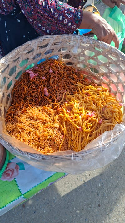 A close-up shot captures a woven basket filled with three different types of noodles, arranged as if for sale. The basket is lined with clear plastic. The noodles range in color from a pale yellow to a dark reddish-brown, with some thinner strands mixed in. Small pink pieces, possibly crackers or spices, are interspersed among the noodles. A person's hand, with visible wrinkles and manicured nails, is partially seen reaching towards the noodles from the right side of the frame. The person is wearing a dark garment with a patterned overlay featuring geometric shapes in shades of red, white, and green. The basket sits on a table covered with a plastic cloth that has a green and white checkered pattern with a rose motif. The background is blurred, showing what appears to be a street scene with pavement and a hint of sunlight creating shadows. The overall lighting suggests it is daytime, possibly outdoors. The location is identified as Grong-Grong, Indonesia.