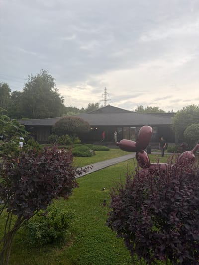 The image shows an outdoor scene in front of a dark-colored building with a pitched roof. The building has large windows and appears to be an entrance with a walkway leading up to it. The walkway is made of light-colored wood planks.
In the foreground, there are several large, dark red, balloon-like sculptures. One is prominently featured on the right side of the image, partially obscuring the building and the walkway. The sculptures are shiny and appear to be made of a smooth, hard material.
Several people are visible near the building entrance. One person in a black shirt stands on the walkway near the top of the stairs. Another person in a white shirt is also visible near the stairs, and a third person in a red shirt is further down. The people appear to be casually dressed.
The setting is outdoors, with a well-maintained green lawn and various bushes and trees surrounding the building and sculptures. The trees are lush and green, indicating it is likely springtime or summer.
The sky is overcast with a bright, diffused light, suggesting it might be daytime, possibly late afternoon or a cloudy morning. There are visible power lines and a pylon in the background, indicating the presence of infrastructure.
The overall mood is calm and perhaps a bit surreal due to the prominent balloon sculptures. The scene seems to be a public space or an art installation outside a building. The location is identified as Zao, Russia. There is no visible text in the image.