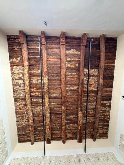 The image shows a close-up, upward-facing view of an old, wooden beamed ceiling with painted decorations. The beams are rough-hewn and appear to be made of dark, aged wood. Between the beams, horizontal wooden planks form the ceiling surface. These planks are decorated with intricate, colorful patterns, primarily in shades of orange, white, and brown, featuring floral motifs and what appear to be stylized figures. Two thin, dark metal rods are suspended vertically from the ceiling, one in front of the second and third beams from the left, and the other in front of the fourth and fifth beams from the left. The surrounding walls are painted a light cream color.
This ceiling is located in the **Palazzo dei Barnabiti in Maddaloni, Italy**, a historic building that served as a monastery and later a public library. The style of the painted beams is characteristic of rural Italian folk art from the 18th or 19th century, often found in aristocratic homes or religious buildings of the period.