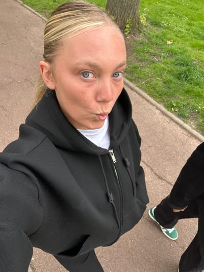 A young, light-skinned woman with blond hair and blue eyes is taking a selfie outdoors. She is wearing a white t-shirt under a black zip-up hoodie and black pants. She has puckered lips. Another person's legs and green sneakers are partially visible in the upper right corner. The ground is paved, and there is grass and a tree visible in the upper left corner. Based on known imagery, the scene could plausibly be located in Noisiel, France.