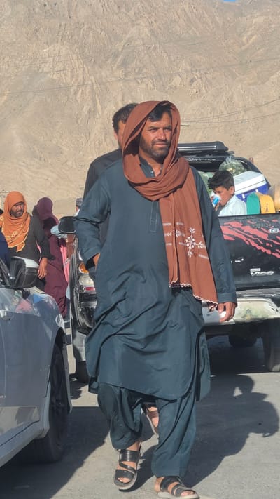 A medium shot shows a group of people crowded on and around a black Toyota Hilux pickup truck in Bostan, Pakistan. The terrain around them is dry and mountainous.

A man in the foreground wears a dark-blue robe and a brown scarf with white embroidery around his head. He is looking at the camera. Behind him, another man is visible, similarly dressed. On top of the truck bed, another man is asleep, lying on top of luggage. Further down, three women are sitting, with two wearing orange and purple hijabs. A silver car is parked nearby. Shadows are visible on the ground, suggesting a sunny day. Powerlines can be seen in the background.