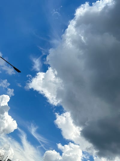 This media file is a photograph of a bright blue sky filled with a variety of clouds. A large, dark gray cloud dominates the upper center of the frame, contrasting with fluffy white cumulus clouds scattered throughout the scene. Thin, wispy cirrus clouds are also visible, adding texture to the sky. In the lower left quadrant, a black lamppost extends upwards into the sky at an angle. The image appears to be taken during the daytime, given the bright sunlight illuminating the clouds. The weather seems to be changeable, with both dark and bright clouds present, suggesting a dynamic atmosphere. The lamppost suggests an outdoor, possibly urban or semi-urban, setting. The image conveys a sense of openness and the vastness of the sky. No people or text are visible.