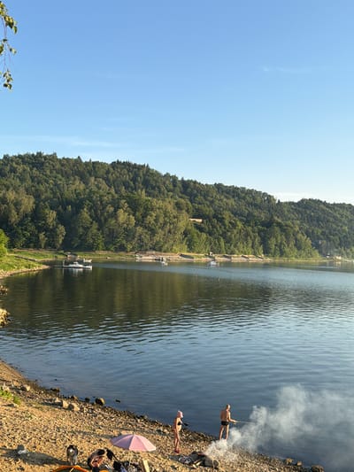 The image depicts a serene lakeside scene in Lipie, Poland. A calm body of water dominates the foreground and middle ground, reflecting the clear blue sky. On the far bank of the lake, a dense, lush green forest covers a hillside that slopes upwards. In the distance, several small boats are moored near the shore, indicating a recreational area.
The immediate foreground shows a gravelly beach. Two individuals are present on the beach. One person, dressed in swimwear, stands by the water's edge, holding a fishing rod and seemingly engaged in fishing. They are surrounded by a significant amount of white smoke, likely emanating from a barbecue or campfire. Another person, also in swimwear, stands a short distance away, observing. Near them, a pink umbrella provides shade over what appears to be camping equipment, including a tent and various other items. A dog, possibly a husky breed, is also visible on the beach, near the tent. The overall atmosphere is one of a peaceful summer day at the lake.