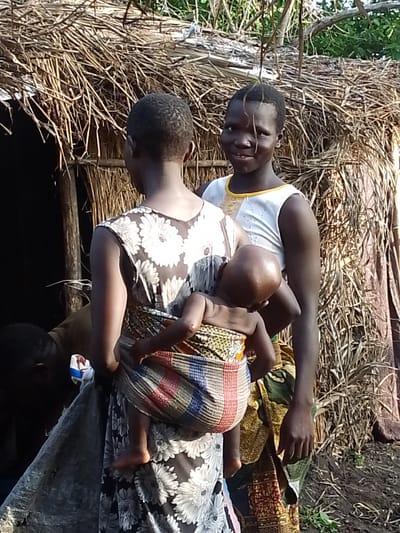 In Kajola, Nigeria, three individuals are depicted in front of a rustic hut constructed from straw and wood. The primary focus is on a woman carrying a baby on her back, wrapped in a colorful cloth. To her right stands another young person, seemingly a girl, looking towards the camera with a slight smile. The hut's entrance is visible to the left, partially obscured by the figures. The ground appears to be dirt, with some sparse vegetation. The overall scene suggests a rural, possibly impoverished, environment.