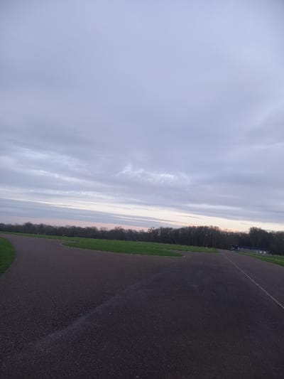 An asphalt track curves along a grassy border separating it from a line of trees. The track dominates the right side of the frame, exhibiting a rough, dark texture. To the left of the grass and trees, the sky takes up the majority of the image, displaying a range of gray and faint orange hues, with streaky clouds extending toward the horizon. The overall lighting appears soft and diffused.