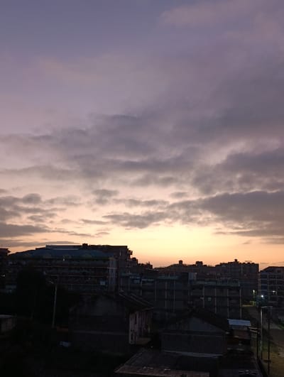The image showcases an urban landscape at dawn or dusk, with multiple buildings silhouetted against a vibrant sky. The sky transitions from a purple hue at the top to a warm orange near the horizon, punctuated by layers of gray clouds. Below, a dense collection of residential structures and apartments are visible, with some buildings carrying the inscription "FORTUNE APARTMENT". The scene evokes a serene yet bustling atmosphere.