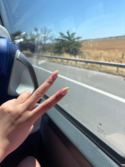 A hand with neon yellow-tipped French manicured nails is held up in front of a window, forming a peace sign. The hand is positioned on the right side of the frame. Through the window, a sunny, rural landscape is visible, with a highway and fields of dry grass or crops. The seat of a vehicle, possibly a bus or train, is visible on the left side of the frame. The window has a dark frame and appears to be tinted. There is some text and a circular marking on the window. The overall impression is a snapshot of a scenic journey on a bright day.