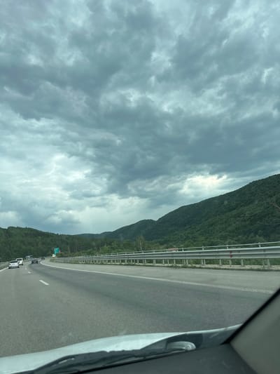 The image depicts a view from inside a vehicle traveling on a highway in Potop, Bulgaria. The highway is asphalt with white lane markings and a metal guardrail separating it from the surrounding environment. On the right side of the highway, a densely forested green hillside rises, dotted with trees and some power line pylons visible near the crest. The sky is overcast with heavy, dark gray clouds, suggesting impending or ongoing rain, with a lighter patch of sky visible in the distance. Several vehicles, including cars and a bus, are visible on the highway in the distance. The foreground shows the hood and windshield of the vehicle from which the photo is taken.