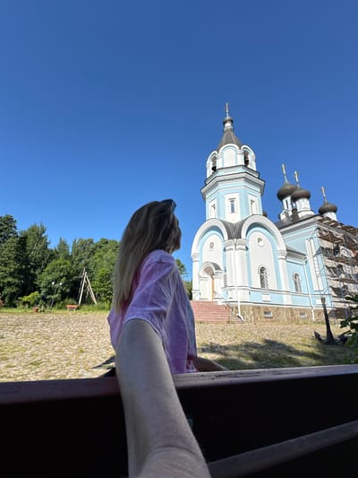 A woman with long blonde hair, wearing a pink tie-dye t-shirt and sunglasses, is standing in front of a light blue and white Orthodox church with multiple domes, located in St. Ozerki, Russia. The church appears to be under renovation, with scaffolding visible on one side. The ground in front of the church is paved with stones, and there are trees and green grass in the background. The sky is clear and blue. The photo is taken from a low angle, with the woman's arm and hand in the foreground, suggesting a selfie or a photo taken by someone close to her.