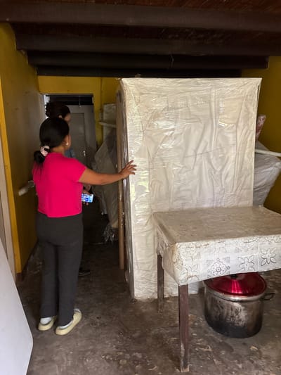 The image shows two people in what appears to be a storage area in Aucallama, Peru. One woman, facing towards the right side of the image, wears a bright pink t-shirt, dark pants, and light-colored shoes. She is holding a smartphone and reaching towards a large, mattress-sized object covered in plastic wrap. Another person is visible in the background near a doorway. The area seems to be indoors with yellow and brown walls. Beside the mattress and woman is a large pot.