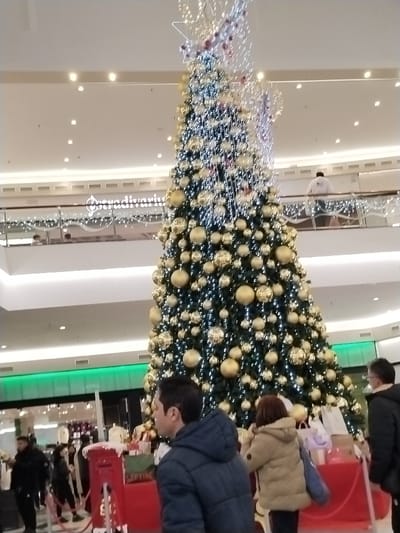An immense, brightly lit Christmas tree adorns a shopping mall. Decorated with golden spheres and sparkling lights, it stretches almost to the roof. A red platform with gifts and a postal box surrounds its base. People mill around the tree, some wearing winter coats. A balcony with a store sign is visible in the background. The scene conveys a festive holiday atmosphere within a modern commercial setting.