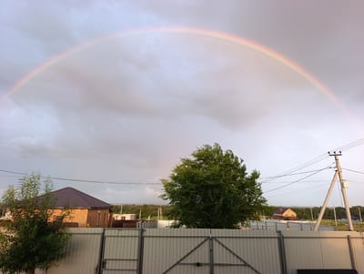 The image depicts a suburban landscape under a sky filled with clouds and a prominent rainbow arching across it. In the foreground, a light-colored metal fence with a double gate runs horizontally across the frame. To the left of the fence, a house with a dark brown roof and brick exterior is visible, partially obscured by a leafy green tree. To the right of the fence, a large, dense green tree stands prominently. Further in the background, several other houses, some with visible roofs, are scattered amongst trees and vegetation. A utility pole with wires stretches across the upper right portion of the image. The overall scene suggests a rural or semi-rural environment in the city of Lenina, Russia, after a rainfall, with the natural phenomenon of a rainbow adding a touch of beauty to the otherwise ordinary setting. There are no people or discernible actions taking place in the image.