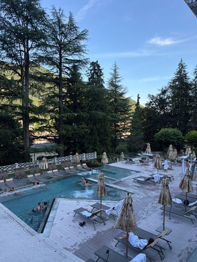 In San Pellegrino Terme, Italy, a resort pool area is depicted under a partly cloudy blue sky. Lush green trees and hills form a backdrop. The pool complex features a main pool with a jacuzzi area and a shallower section, both filled with clear blue water. Several people are visible in the water, including couples and individuals. The surrounding deck is furnished with numerous lounge chairs, many of which are unoccupied, and decorative umbrellas with floral patterns. A white lattice fence separates the pool area from a row of lounge chairs further back. Two individuals in white bathrobes stand near each other on the deck, while another person in a bathrobe reclines on a lounge chair. The scene conveys a relaxed atmosphere at an upscale outdoor spa.