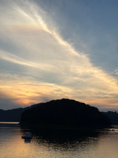 A serene sunset over Lake Rożnowskie in Gródek nad Dunajcem, Poland, captures a tranquil scene. The sky is painted with a gradient of soft orange and yellow hues near the horizon, transitioning to a pale blue at the top, streaked with delicate, wispy clouds. In the foreground, the still waters of the lake reflect the warm colors of the sunset, creating shimmering patterns. A small, white floating structure, possibly a buoy or a small hut, sits on the water to the left, its reflection clearly visible beneath it. Silhouetted against the vibrant sky, a large, tree-covered island dominates the middle ground, its dark outline sharp and defined. In the distance on the right, a small boat with two occupants can be seen navigating the water. The surrounding hills on the far bank are also silhouetted in dark tones, creating a dramatic backdrop to the illuminated sky and water. The overall impression is one of peace and natural beauty.
