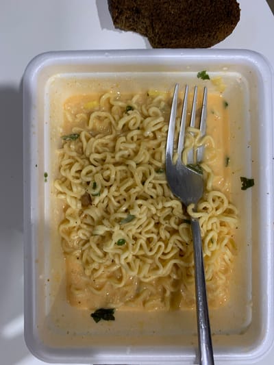 A top-down, medium shot captures a white, rectangular, disposable food container filled with instant ramen noodles. The noodles are yellowish and curly, submerged in a creamy, orange broth with visible flecks of green herbs and other seasonings. A silver fork with four tines is positioned in the right portion of the container, with some noodles wrapped around its prongs, indicating that the meal is partially consumed. In the upper right corner of the frame, a dark, irregularly shaped piece of bread or toast is visible, positioned on a white surface. The scene appears to be indoors, likely on a table, with a plain white background that extends to the left and top of the container. The lighting suggests an overhead light source, casting subtle shadows. There is no indication of the city of Ильинское-Усово, Russia.