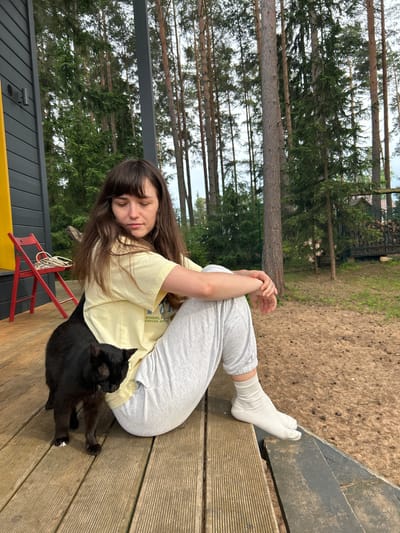 In this media file, a young woman with long brown hair and bangs, wearing a light yellow t-shirt and grey sweatpants with white socks, is relaxing on a wooden deck. She is curled up, with her eyes closed and her head resting on her arm, appearing to be asleep or deep in thought. A black cat is nestled beside her, also seemingly at rest. The scene is set outdoors, amidst a backdrop of tall pine trees, suggesting a natural, possibly wooded, location. The wooden deck, some steps, and a red foldable chair are visible, hinting at a casual outdoor living space, possibly a porch or patio of a house or cabin. The ground beyond the deck is covered with sandy soil and small pebbles, further reinforcing the natural setting. The lighting suggests it is daytime, with soft natural light filtering through the trees. The overall mood is one of tranquility and peacefulness. There is no visible text in the image. The location context is Shepelyovo, Russia.