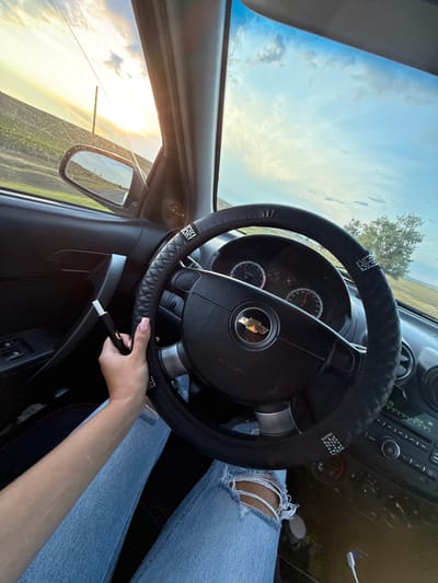 A POV shot from the interior of a car shows a person's left hand holding a dark electronic cigarette device with a white tip near the steering wheel. The person is wearing light blue ripped jeans and is seated in the driver's seat. The car's steering wheel is black with a textured grip and is adorned with rhinestones. The dashboard has a Chevrolet emblem in the center of the steering wheel and displays a speedometer and tachometer. The car's window offers a view of a sunset with clouds in a blue sky, and a rural landscape with fields and a distant tree. The image is taken during golden hour. The car appears to be located on a road in the vicinity of Iordanovca, Moldova, based on the provided context.