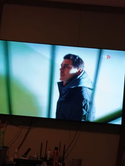 The media file displays a television screen showing a man from the chest up, in profile, facing to the left. He has dark hair and is wearing a dark blue quilted jacket with a high collar. The background on the screen is a bright, somewhat abstract scene with cool, cool-toned lighting, primarily light blue and green hues, suggesting an indoor or studio setting, possibly with artificial lighting. In the upper right corner of the screen, a small logo is visible, reading "TBH" inside a red circle. This logo indicates the television channel broadcasting the program. The overall mood conveyed by the man's expression is serious or pensive. The image of the television is captured from a slightly angled perspective, with the bottom portion of the frame showing a dimly lit area with various electronic devices, cables, and some bottles, indicating the viewing environment. The time of day cannot be determined.