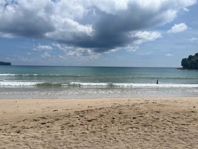 A wide, sandy beach in Ban Bang Thao, Thailand, stretches from the foreground to the ocean. The sand is a light brown color with visible footprints and some scattered debris. Gentle waves roll onto the shore, creating a foamy white line where they meet the sand. The ocean water is a clear turquoise color. In the distance, a single person is standing in the water, appearing as a dark silhouette against the waves. The horizon is marked by the meeting of the sea and sky, which is filled with dramatic, dark gray clouds mixed with patches of blue sky and smaller white clouds. To the far right, a rocky, tree-covered headland juts out into the sea. To the far left, a faint landmass with some structures can be seen in the distance.