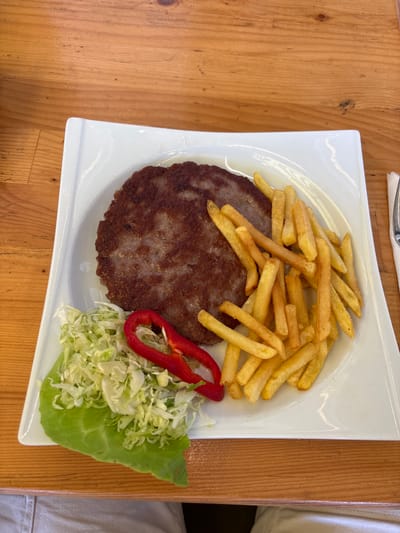 The image depicts a white square plate containing a ground meat patty, a pile of french fries, shredded cabbage, and a slice of red pepper. The plate rests on a wooden surface. The image was taken in the city of Velika Plana Municipality, Serbia. No people are visible in the frame. The interaction is implied, with the food presented for consumption. The environment is a simple table setting.